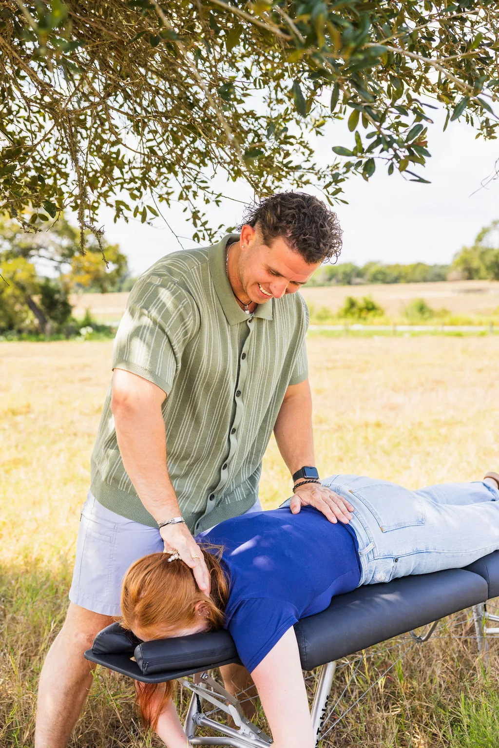 Dr. Levi Hansen examines a patient lying prone on a portable chiropractic table outdoors under tree shade.