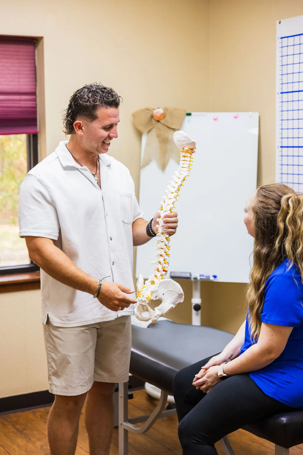Dr. Levi Hansen holds an anatomical spine model while speaking with a patient seated on a treatment table.
