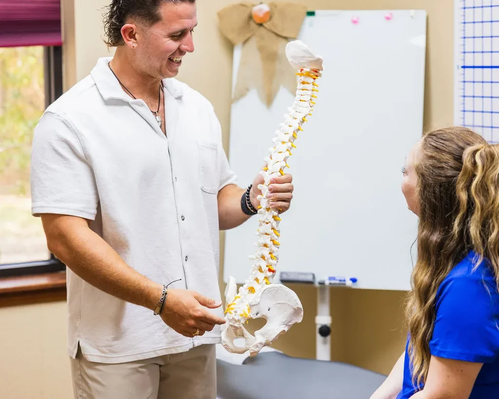 Dr. Levi Hansen holds an anatomical spine model while speaking with a patient seated on a treatment table.